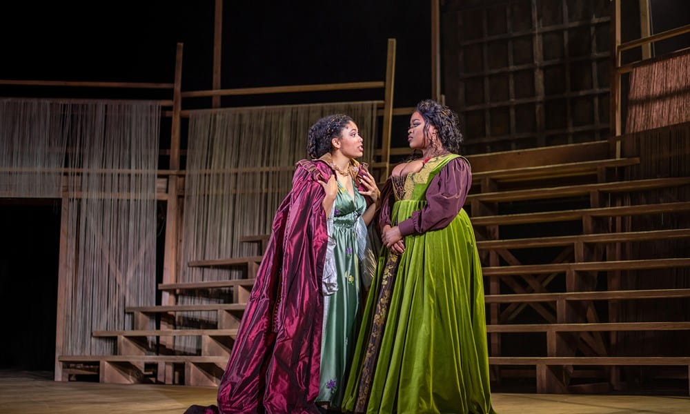 Two women in elaborate period costumes stand on a wooden stage set, engaged in conversation. One wears a red cloak, the other a green dress.