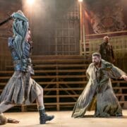 Two actors in medieval costumes engage in a dramatic sword fight on stage, with one person lying on the ground and a wooden set in the background.