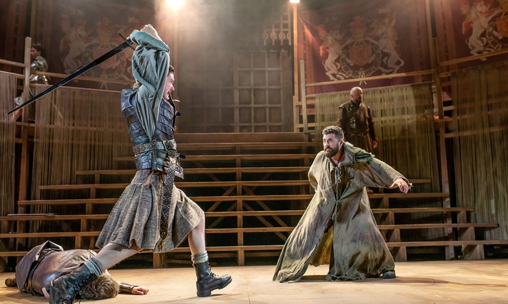 Two actors in medieval costumes engage in a dramatic sword fight on stage, with one person lying on the ground and a wooden set in the background.