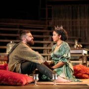 A man in rustic clothing and a woman in a crown and green dress sit on cushions on a theater stage, facing each other, with props and wooden structures in the background.
