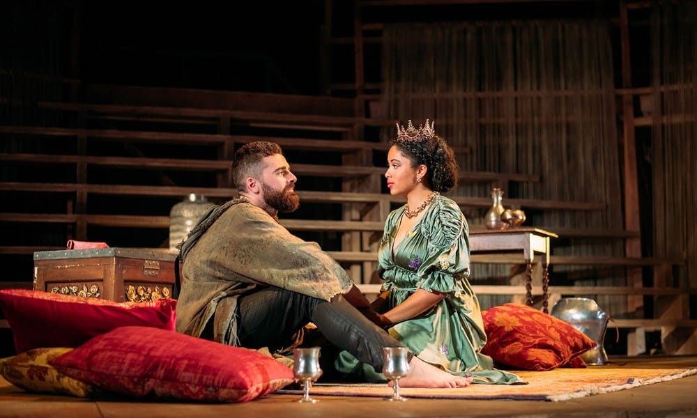 A man in rustic clothing and a woman in a crown and green dress sit on cushions on a theater stage, facing each other, with props and wooden structures in the background.