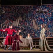 Actors in medieval costumes perform a dramatic scene on stage, with a tapestry featuring unicorns in the background and a banquet table at the center.