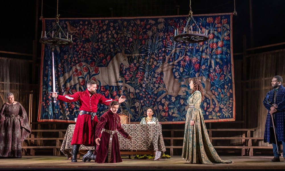Actors in medieval costumes perform a dramatic scene on stage, with a tapestry featuring unicorns in the background and a banquet table at the center.
