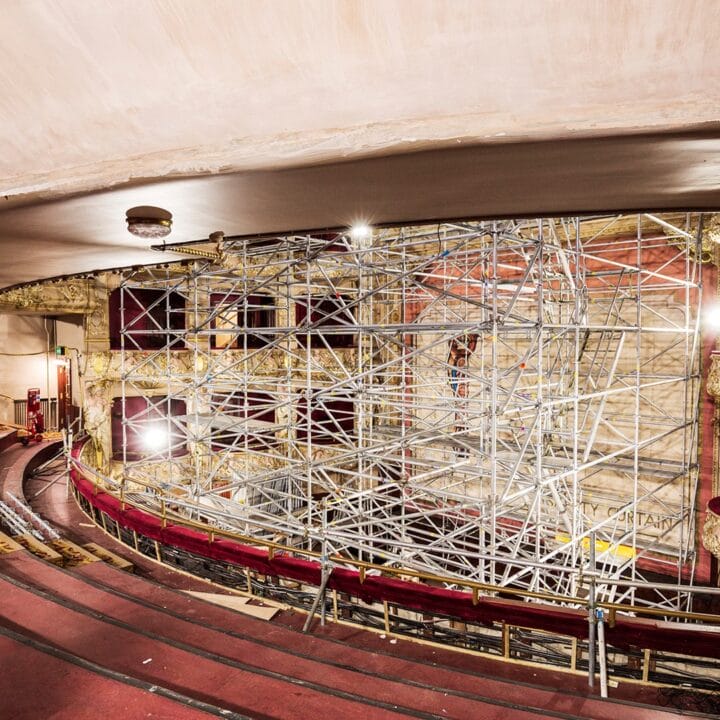 A theater interior with ornate balconies and rows of seats, featuring extensive scaffolding on the stage area for construction or restoration work.