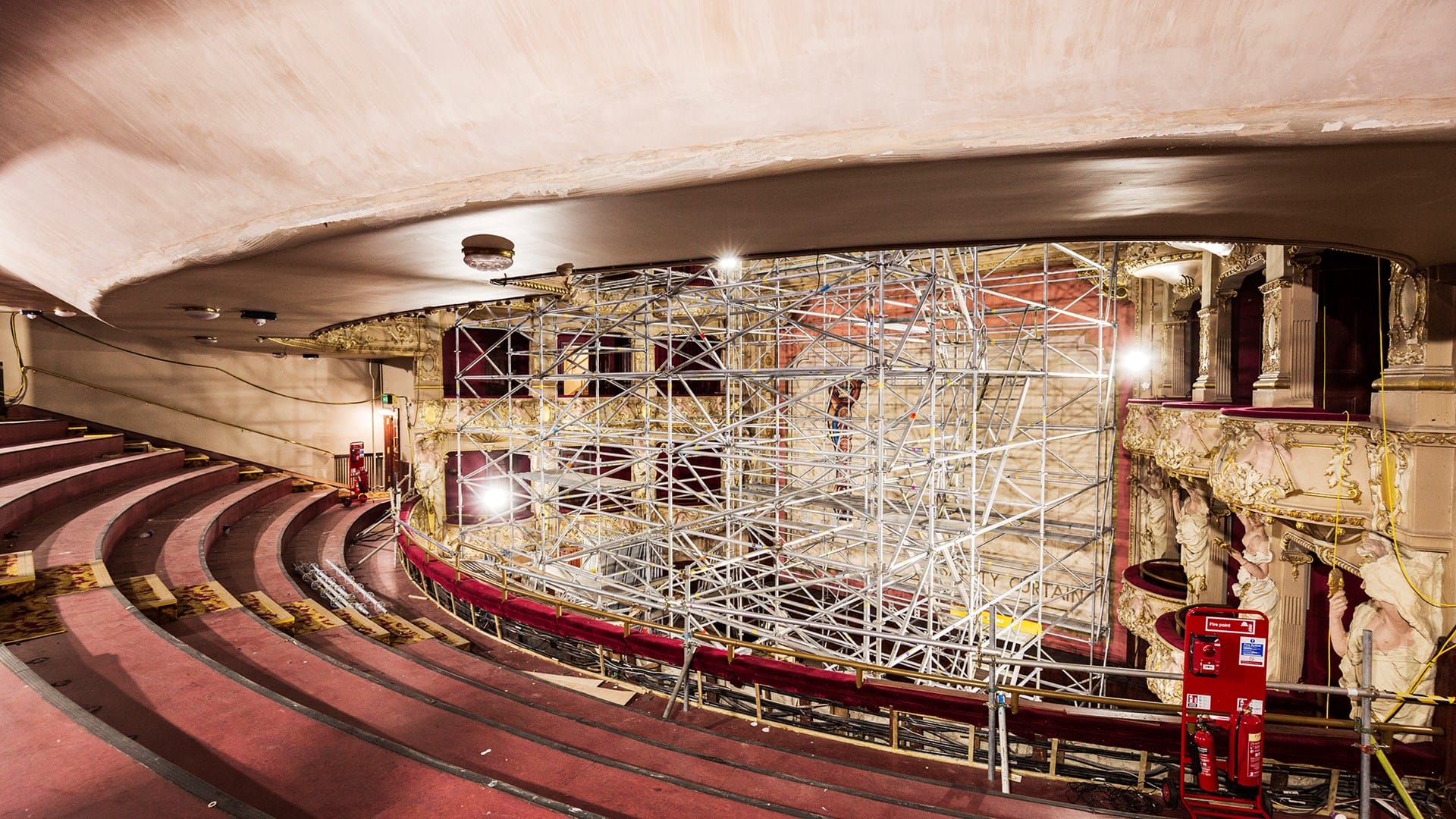 A theater interior with ornate balconies and rows of seats, featuring extensive scaffolding on the stage area for construction or restoration work.