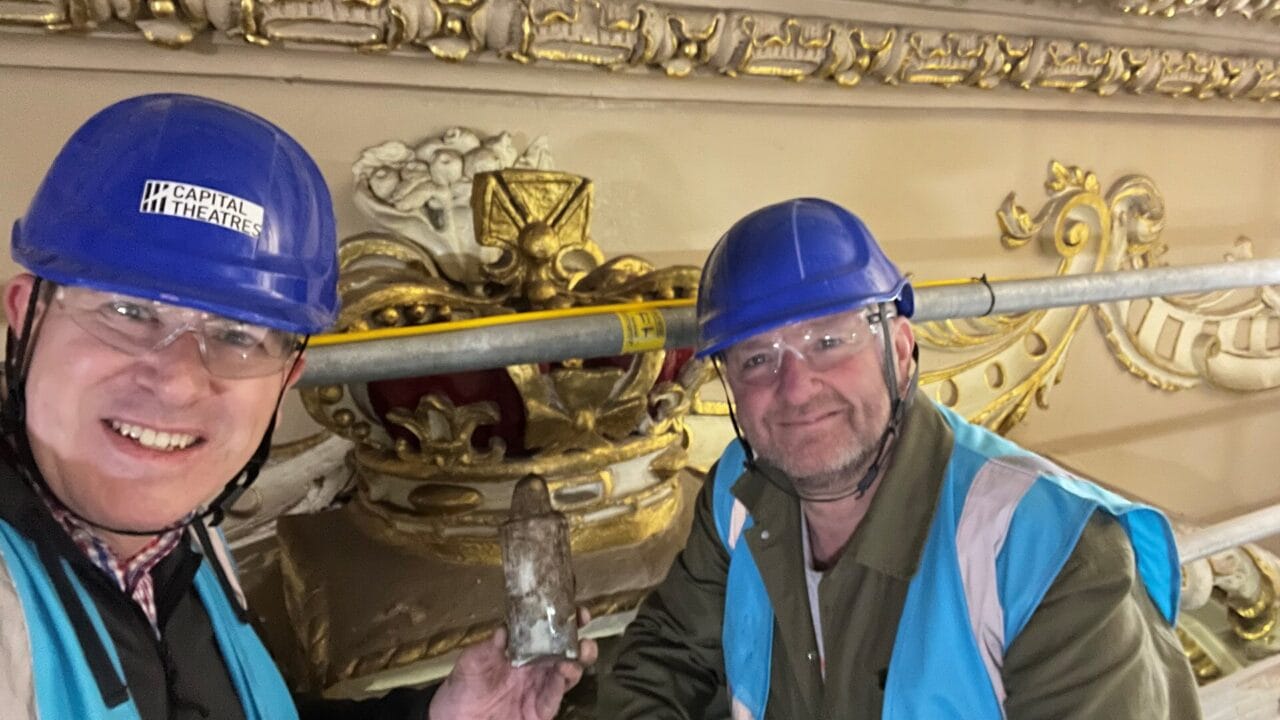 Two men in blue hard hats and safety vests pose with a bottle in front of an ornate gold crown decoration, with scaffolding visible behind them.