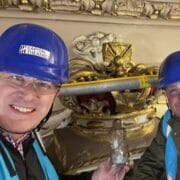 Two men in blue hard hats and safety vests pose with a bottle in front of an ornate gold crown decoration, with scaffolding visible behind them.