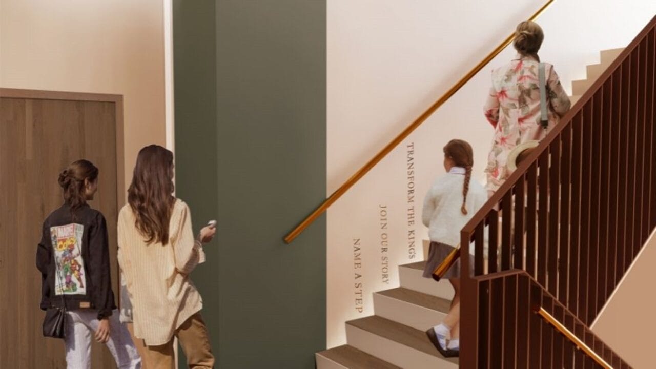 Three women walk toward and one woman ascends a staircase; text on the stair risers reads “Transform the Rings, Join Our Story, Name a Step.”.