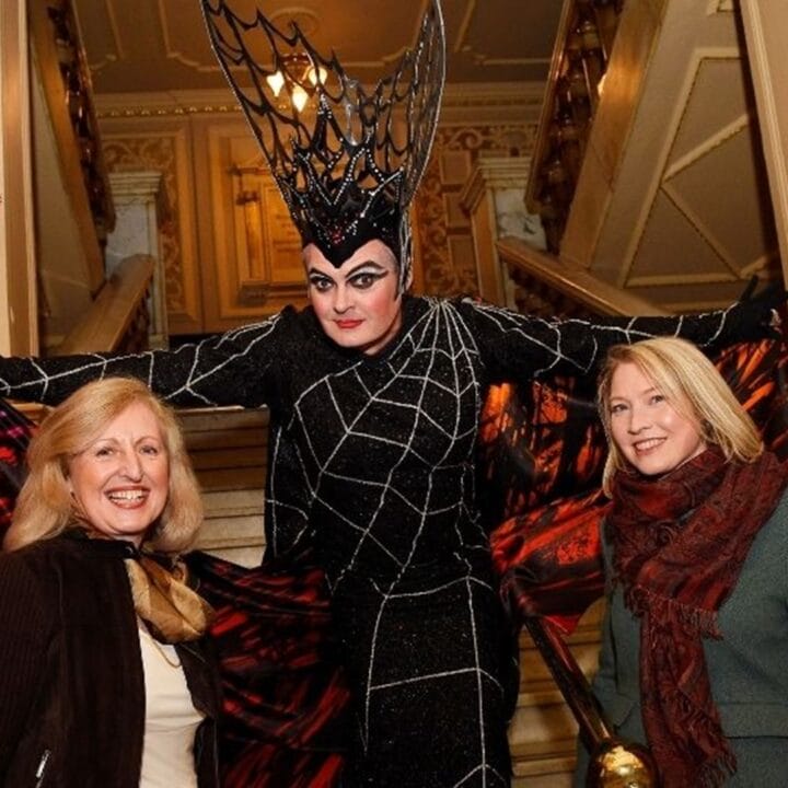Three people pose in front of a staircase; the center person is in a black spiderweb costume with a large headpiece, while the two others wear winter clothing and smile.