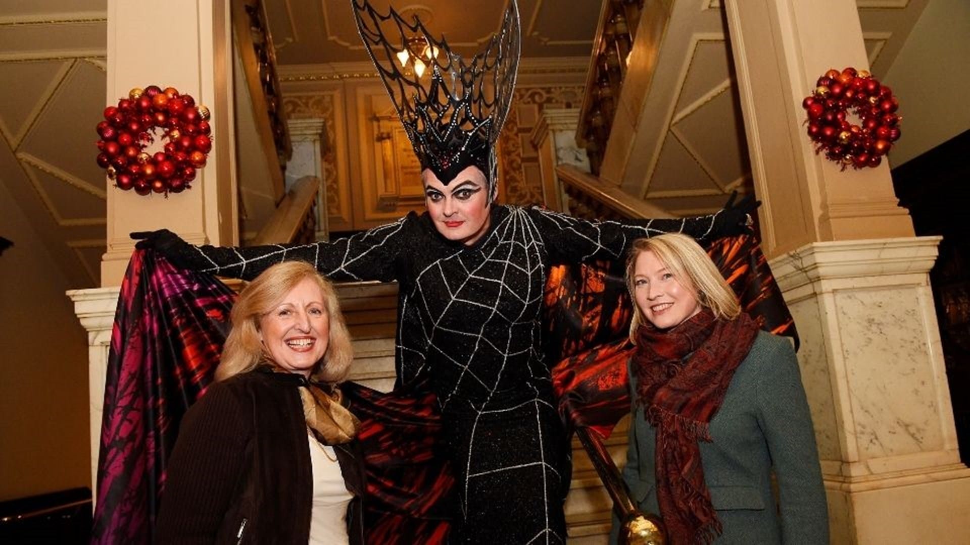 Three people pose in front of a staircase; the center person is in a black spiderweb costume with a large headpiece, while the two others wear winter clothing and smile.