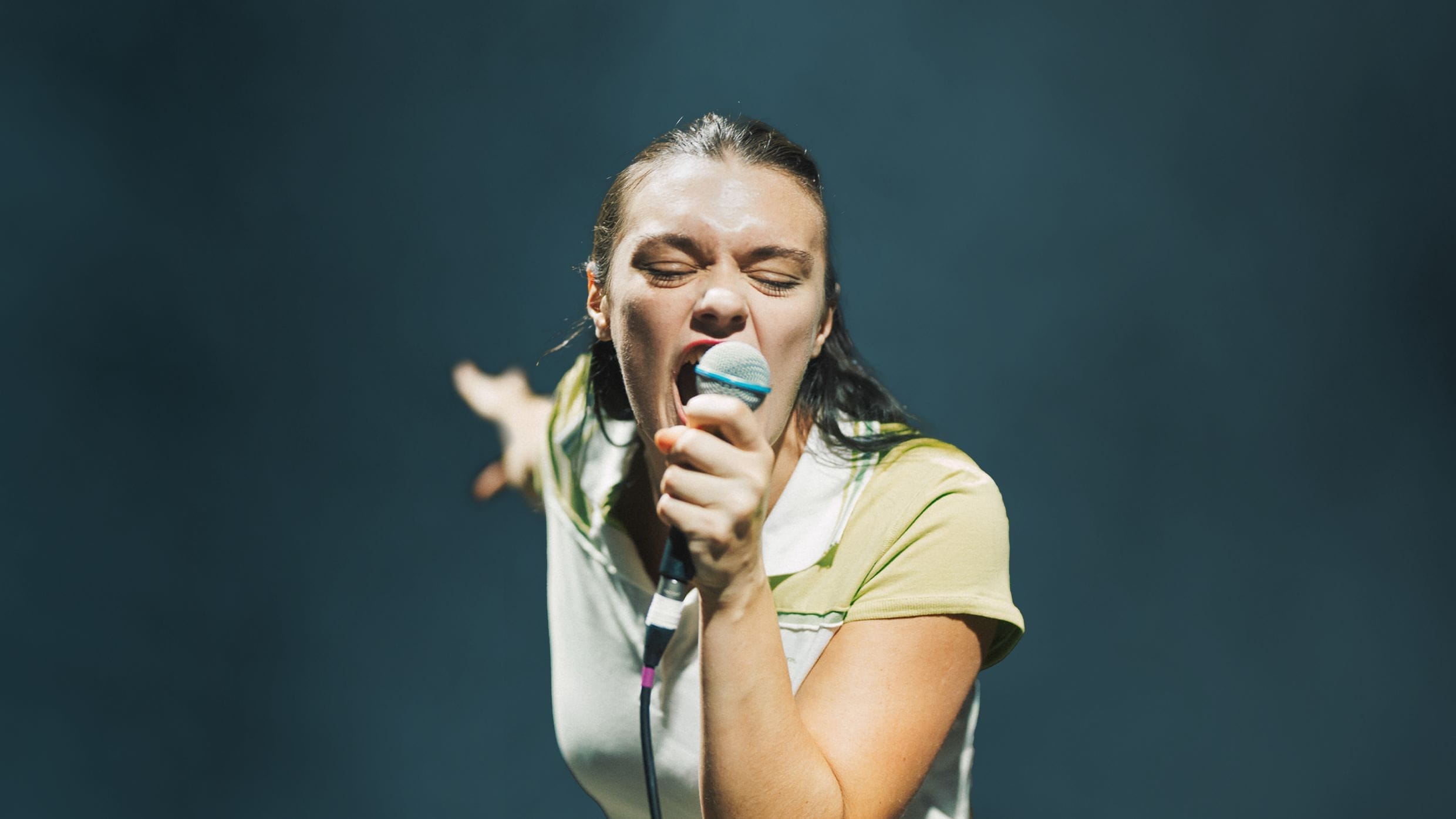 Person with wet hair passionately singing into a microphone on stage, arm outstretched, as Common Tongue lyrics echo against a dark background.