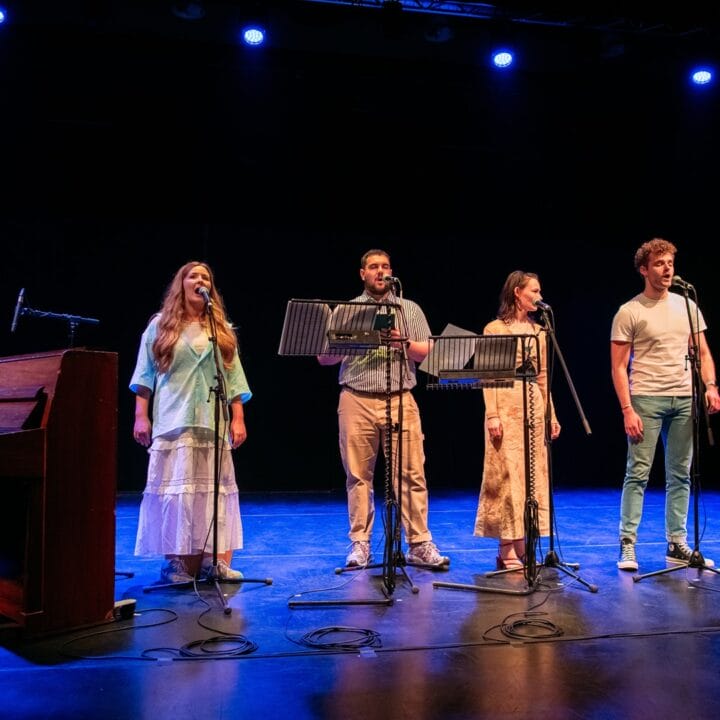 Six performers stand on stage with microphones while a pianist plays on the left. They are under stage lights in a dark theater setting.