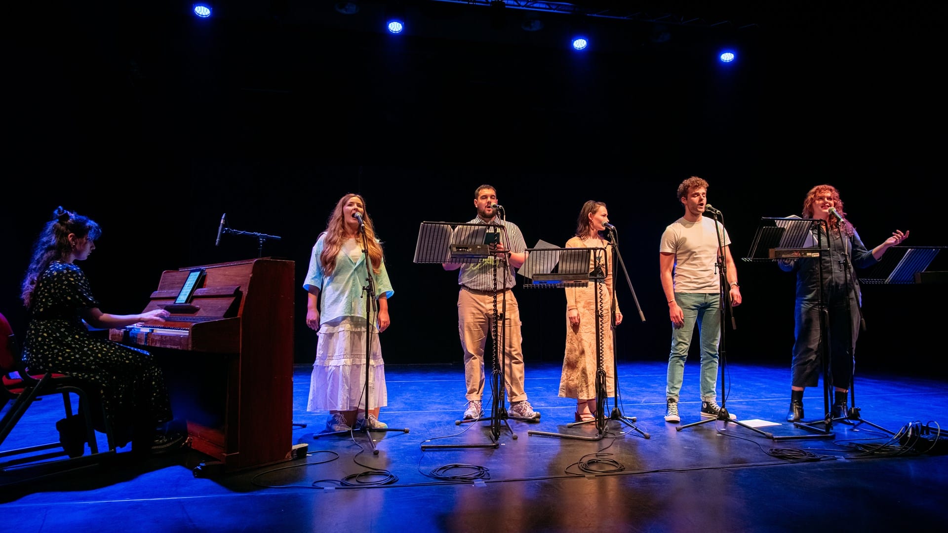 Six performers stand on stage with microphones while a pianist plays on the left. They are under stage lights in a dark theater setting.