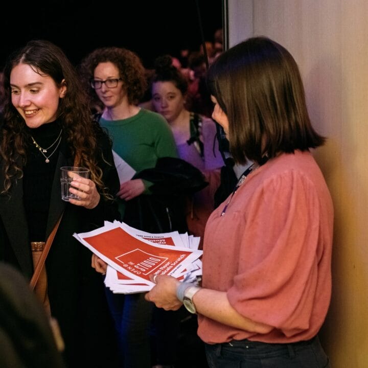 A group of people gathers indoors as a woman hands out red and white flyers; one woman in focus holds a drink and smiles.