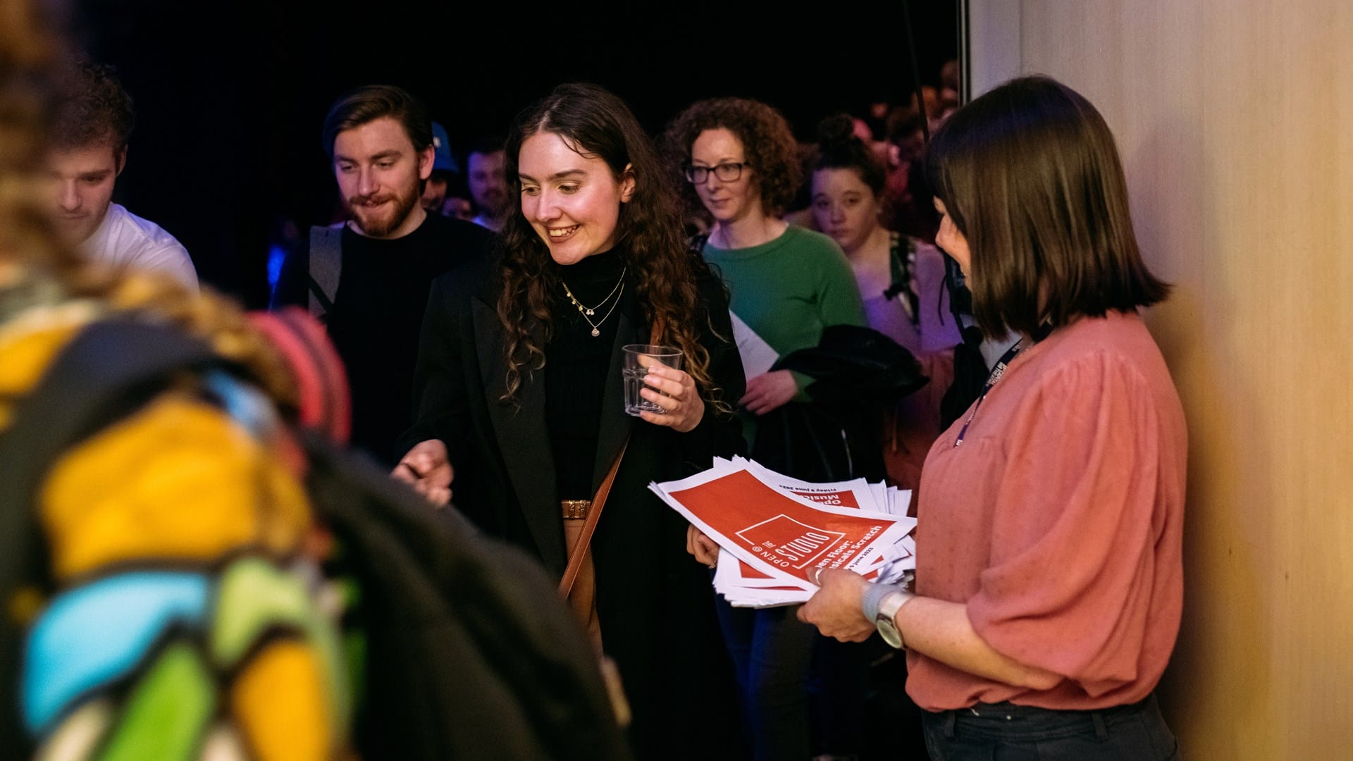 A group of people gathers indoors as a woman hands out red and white flyers; one woman in focus holds a drink and smiles.