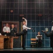 Four actors on stage perform a comedic scene in an office setting, with one actor seated at a desk and the others dancing in mid-step; a large portrait hangs on the wall behind them.
