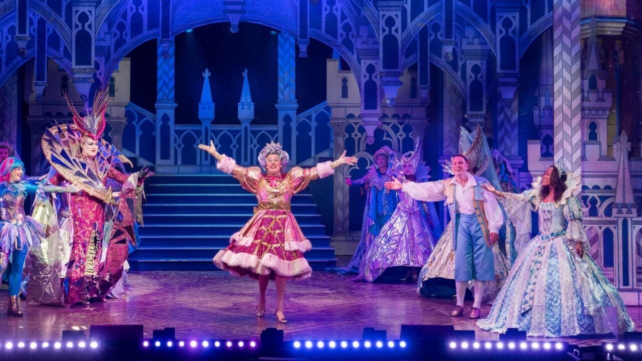 Actors in elaborate, colorful costumes perform on a brightly lit stage designed to look like a palace, with one performer in pink standing center stage, arms outstretched.