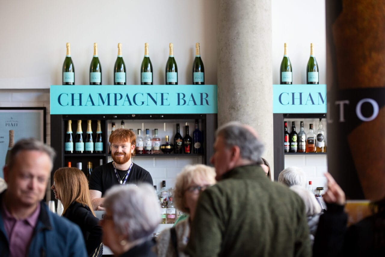 A bartender stands behind the champagne bar, with bottles displayed above and a crowd of people mingling in the foreground.
