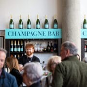 A bartender stands behind the champagne bar, with bottles displayed above and a crowd of people mingling in the foreground.