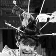 A person wearing a whimsical hat topped with small laundry items and clothespins poses and smiles for the camera.