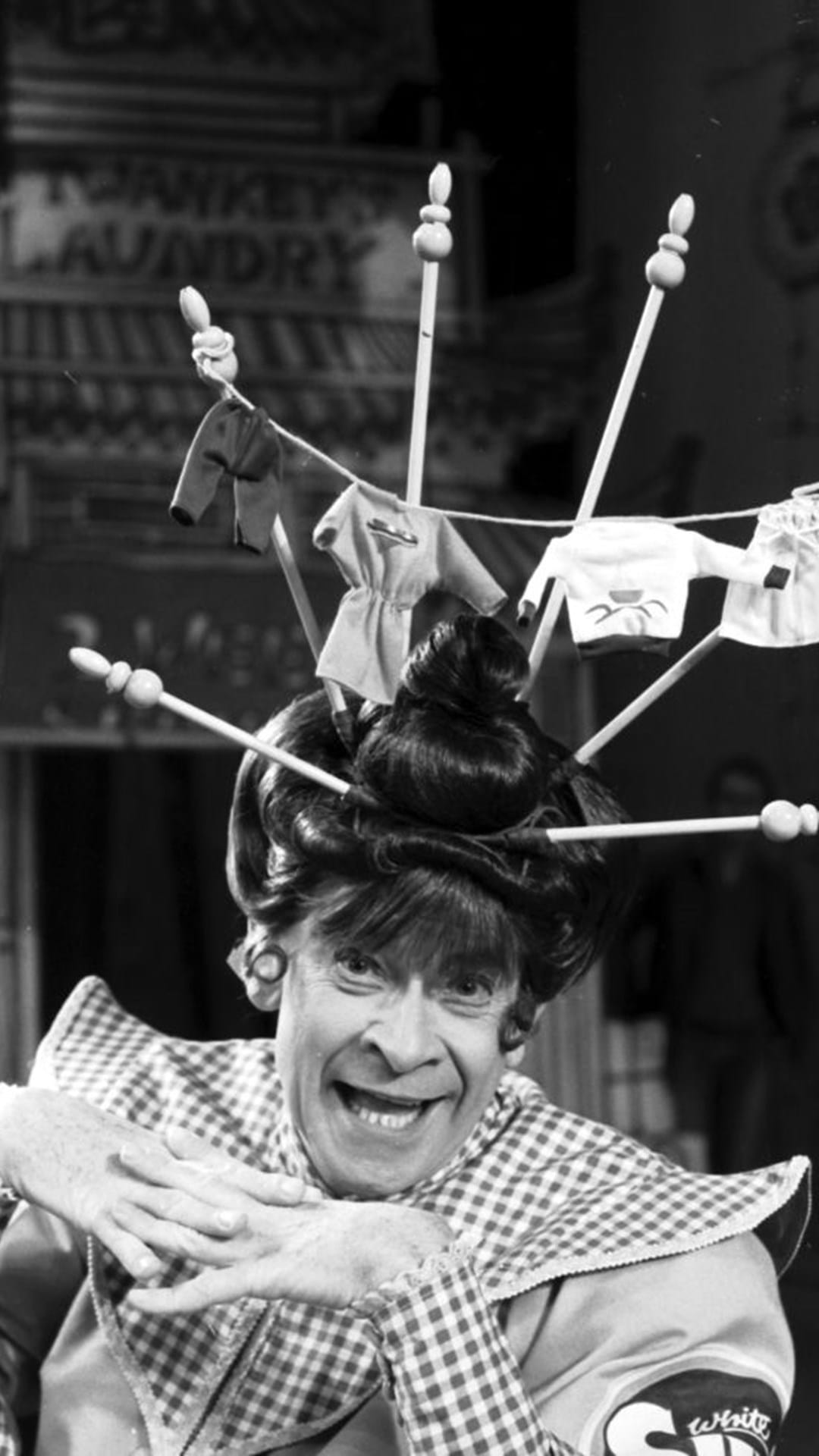 A person wearing a whimsical hat topped with small laundry items and clothespins poses and smiles for the camera.