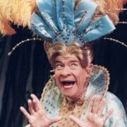 A person in an ornate costume with a large feathered headdress poses with an expressive face and hands raised, wearing a blue and gold embellished outfit.
