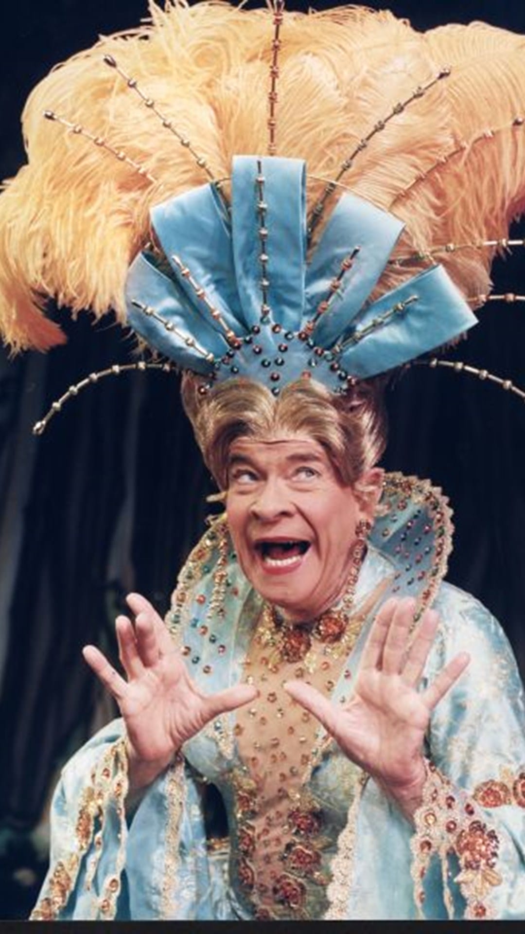 A person in an ornate costume with a large feathered headdress poses with an expressive face and hands raised, wearing a blue and gold embellished outfit.