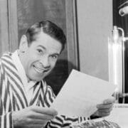 A man in a striped robe sits at a desk under bright lights, smiling while holding a letter and looking toward the camera.