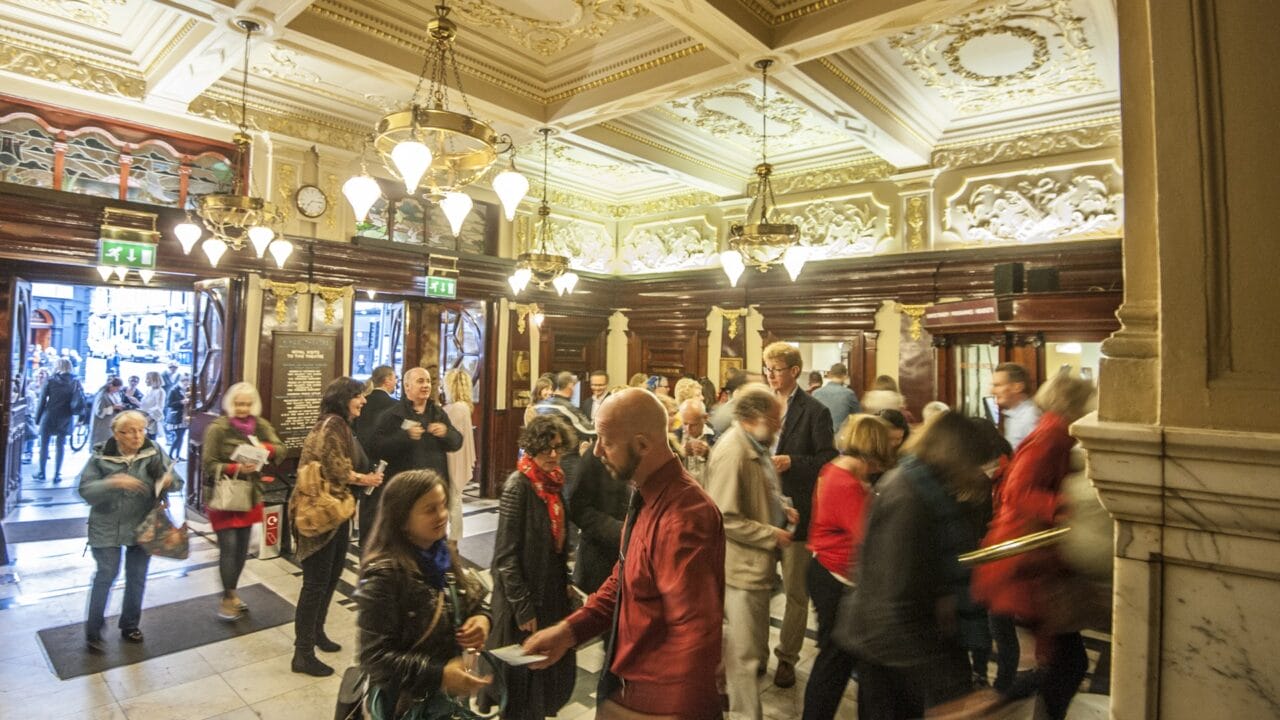 A crowd of people enter a grand, ornate lobby with chandeliers and decorative ceilings, some interacting, while light streams in from open doors.