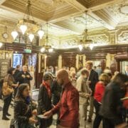 A crowd of people enter a grand, ornate lobby with chandeliers and decorative ceilings, some interacting, while light streams in from open doors.
