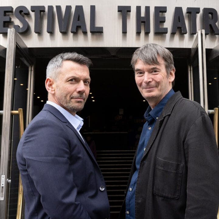 Two men stand outside the entrance of Festival Theatre, facing the camera with the theatre's sign visible above the doors.