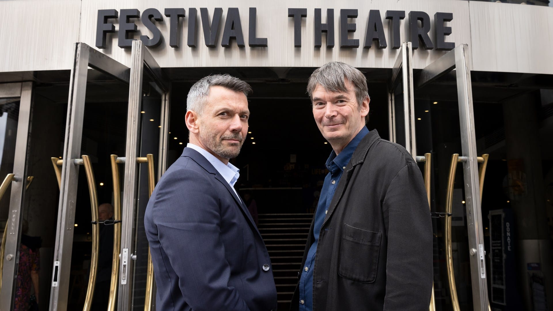 Two men stand outside the entrance of Festival Theatre, facing the camera with the theatre's sign visible above the doors.