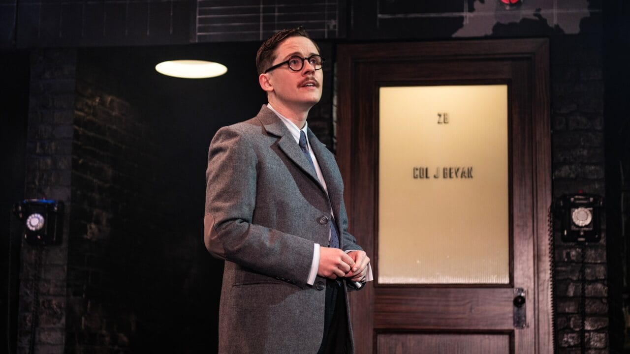 A person in a grey suit and glasses stands in front of a wooden door with frosted glass, featuring black brick walls and a round ceiling light in the background.
