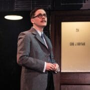 A person in a grey suit and glasses stands in front of a wooden door with frosted glass, featuring black brick walls and a round ceiling light in the background.