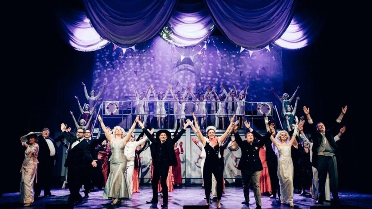 A large group of performers in formal and vintage costumes stand on stage with arms raised, under dramatic lighting and draped curtains, during a theatrical production.