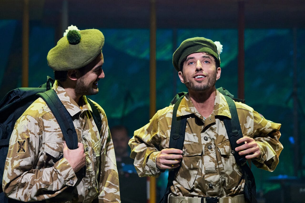 Two men dressed in camouflage military uniforms and green berets stand side by side with backpacks, looking at each other and upward on a stage.