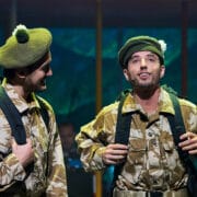 Two men dressed in camouflage military uniforms and green berets stand side by side with backpacks, looking at each other and upward on a stage.