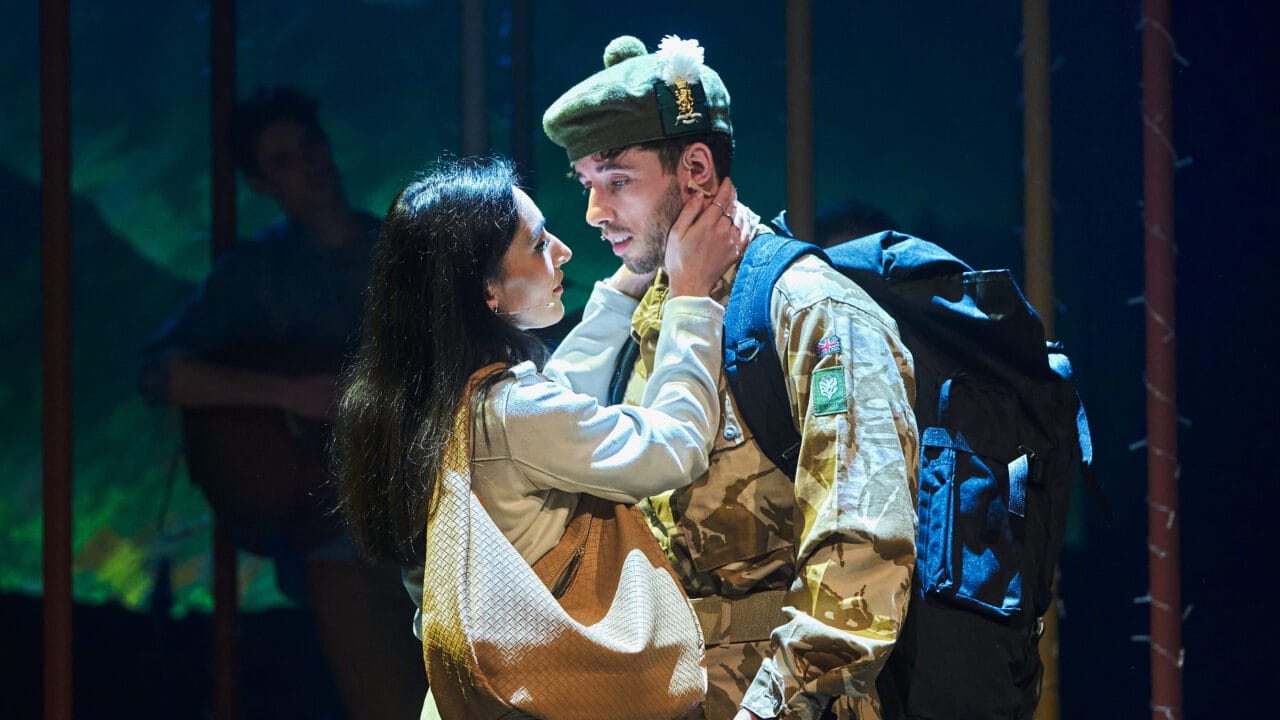 A woman embraces a man in military uniform with a large backpack under stage lighting, suggesting an emotional moment in a theatrical performance.