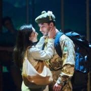A woman embraces a man in military uniform with a large backpack under stage lighting, suggesting an emotional moment in a theatrical performance.