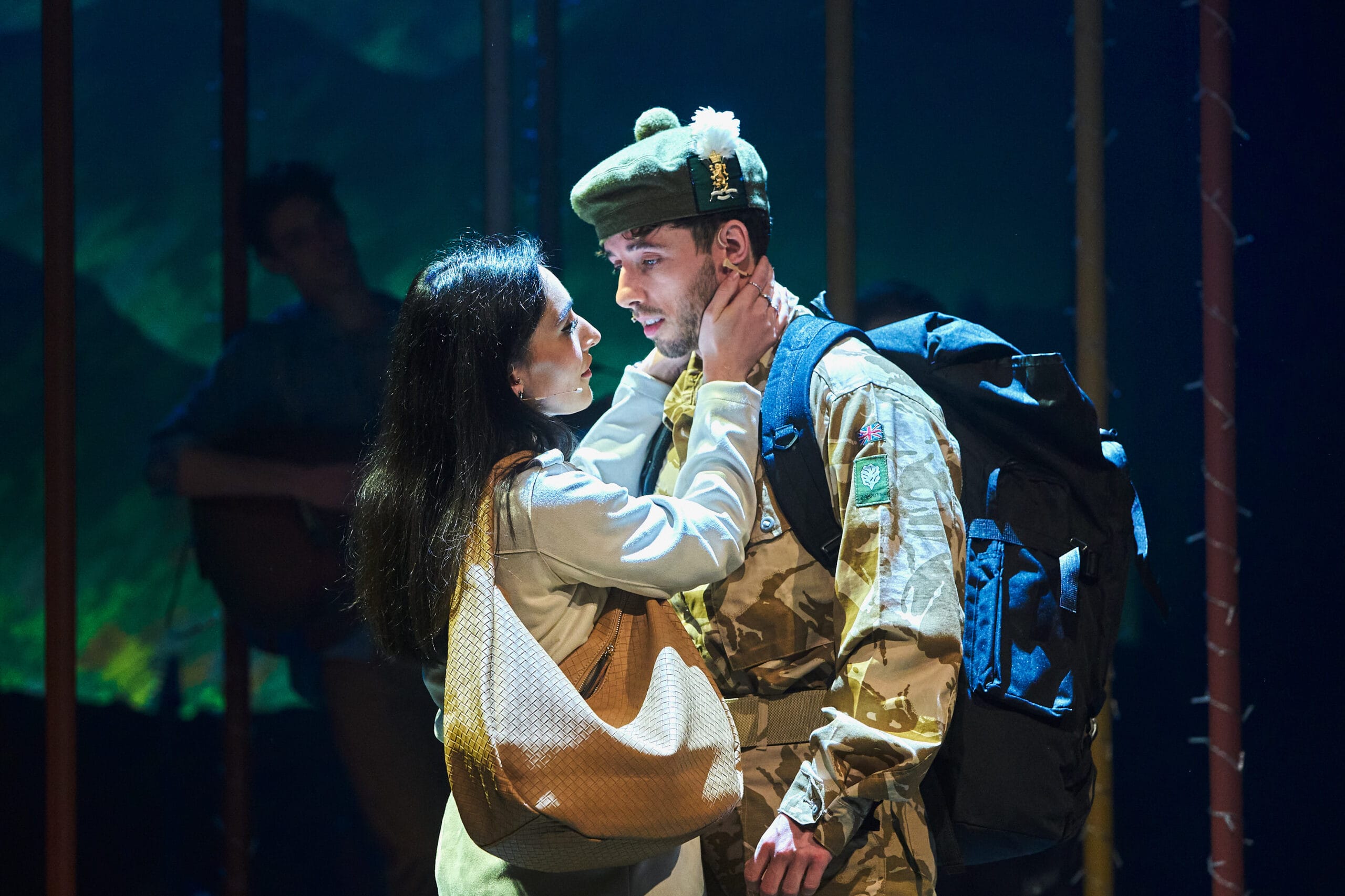 A woman embraces a man in military uniform with a large backpack under stage lighting, suggesting an emotional moment in a theatrical performance.