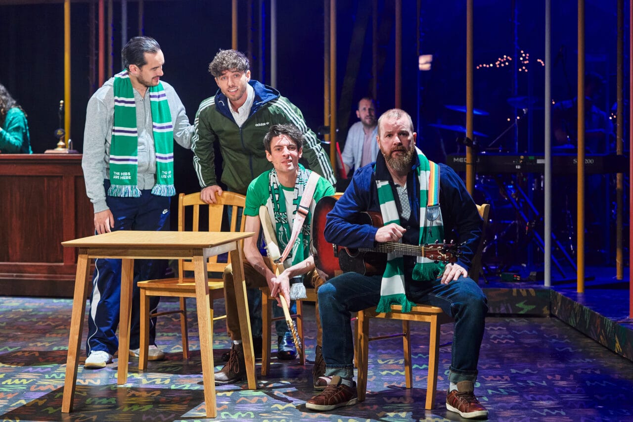 Four men wearing green and white scarves are gathered around a table on stage, with one man seated holding a guitar, suggesting a scene from a theatre performance.