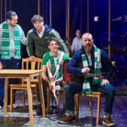 Four men wearing green and white scarves are gathered around a table on stage, with one man seated holding a guitar, suggesting a scene from a theatre performance.