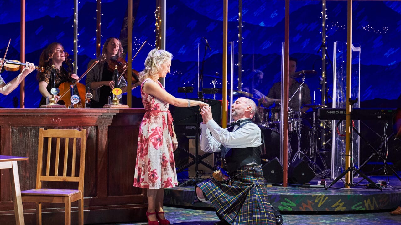 A man in a kilt kneels, holding hands with a woman in a floral dress, onstage beside a bar set, with musicians playing instruments in the background.