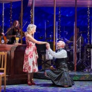 A man in a kilt kneels, holding hands with a woman in a floral dress, onstage beside a bar set, with musicians playing instruments in the background.