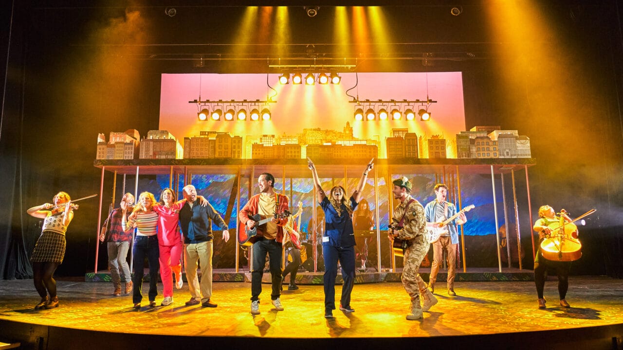A group of performers sing and play instruments on a brightly lit theater stage with a cityscape backdrop.