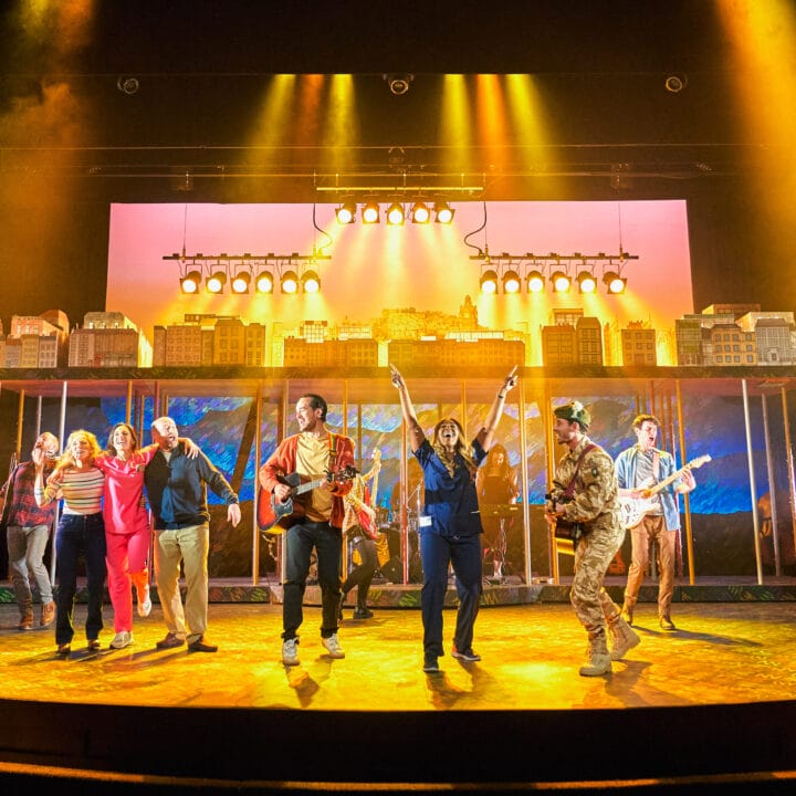A group of performers sing and play instruments on a brightly lit theater stage with a cityscape backdrop.