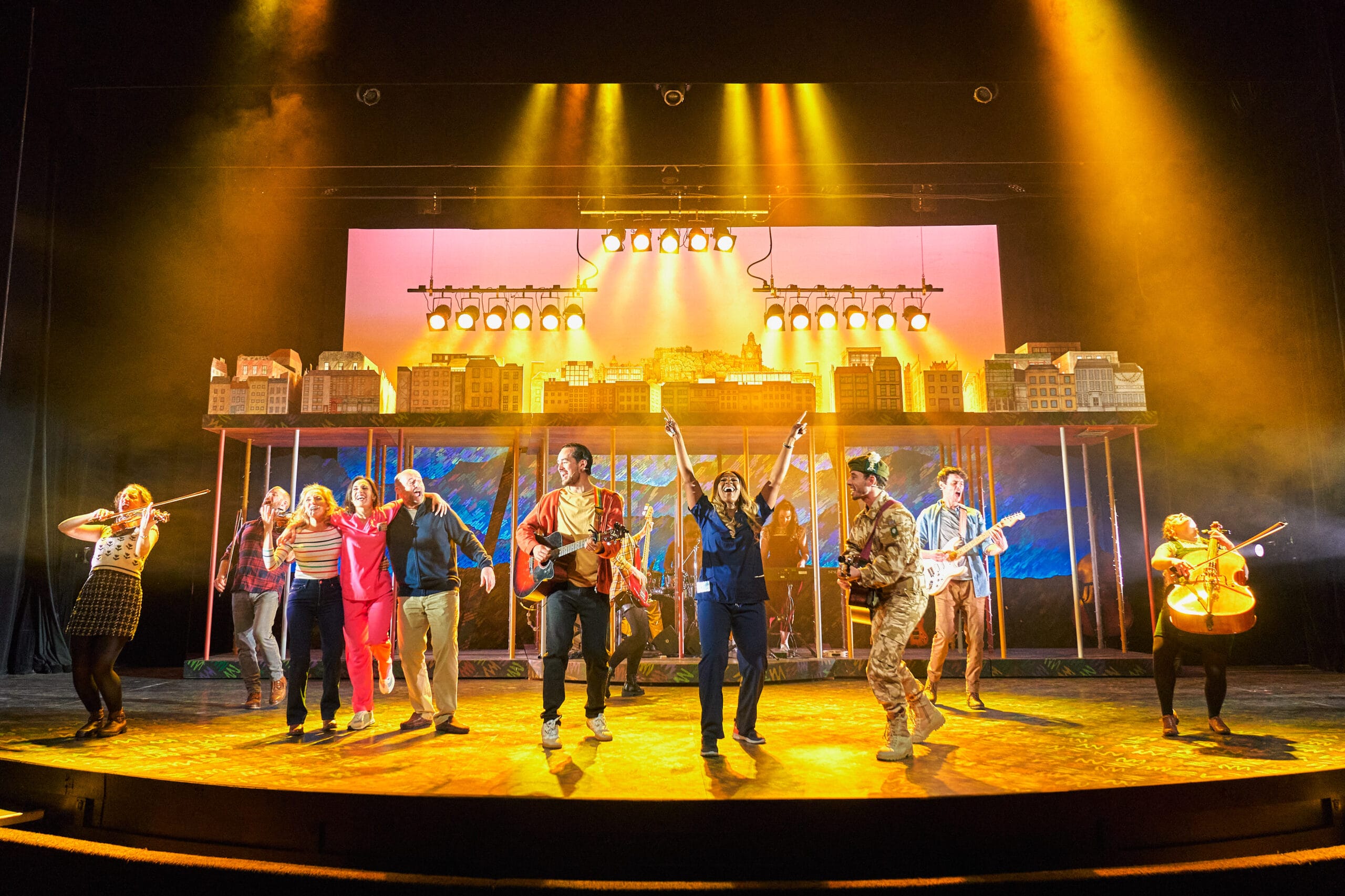 A group of performers sing and play instruments on a brightly lit theater stage with a cityscape backdrop.