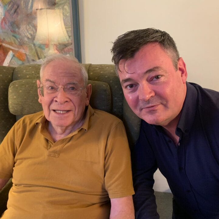 An elderly man in a mustard polo shirt sits in an armchair next to a middle-aged man in a dark shirt; both are looking at the camera indoors.
