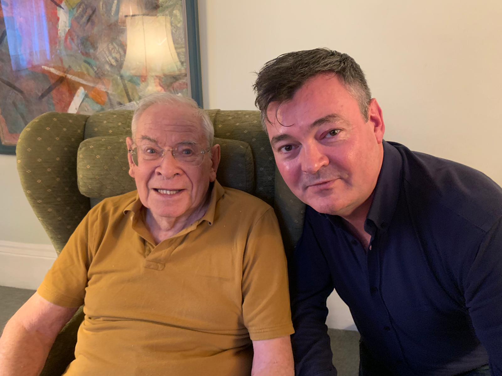 An elderly man in a mustard polo shirt sits in an armchair next to a middle-aged man in a dark shirt; both are looking at the camera indoors.
