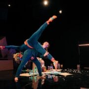 Two dancers perform a balanced acrobatic pose onstage, with one lifting the other. The scene is set in dim lighting with props, a crescent moon, and small lights in the background.